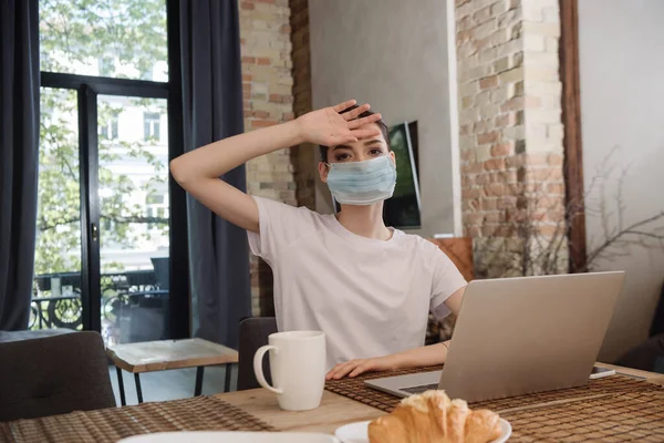 Молодой фрилансер в медицинской маске касается лба рядом с ноутбуком — стоковое фото
