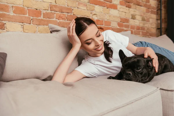Счастливая девушка лежит на диване и трогает черного французского бульдога — стоковое фото