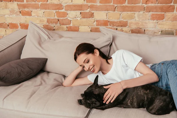 Улыбающаяся девушка лежит на диване и трогает черного французского бульдога — стоковое фото