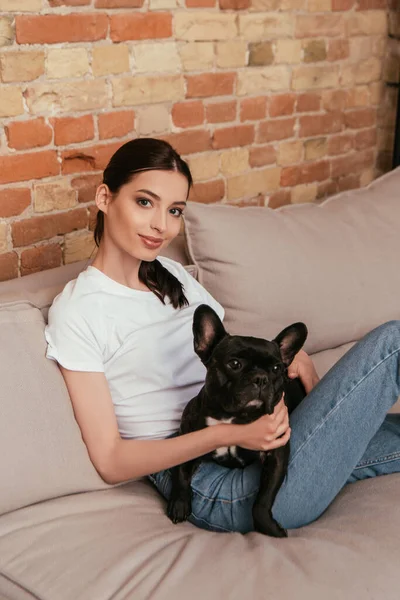 Happy young woman sitting on sofa with black french bulldog — Stock Photo