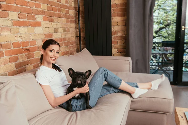 Веселая молодая женщина сидит на диване с черным французским бульдогом — стоковое фото