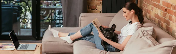 Panoramic crop of happy young woman reading book near black french bulldog and laptop with blank screen — Stock Photo