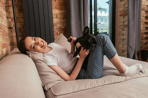 Mujer feliz tocando lindo bulldog francés y mirando a la cámara en la sala de estar - foto de stock