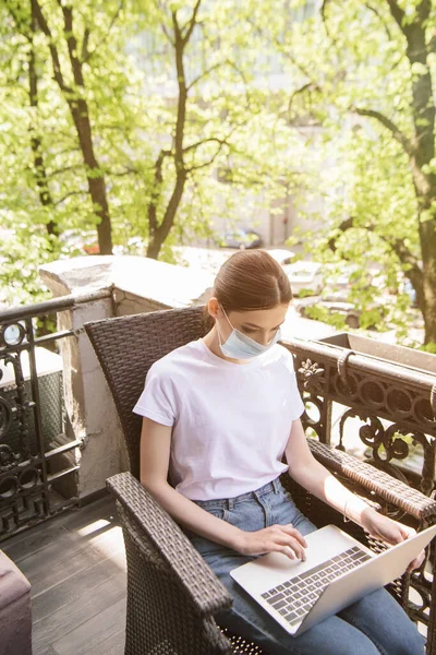 Young freelancer in blue medical mask sitting on chair and using laptop on balcony — Stock Photo