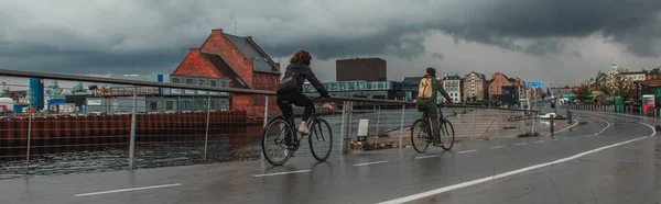Cultivo panorámico de personas en bicicleta en la calle urbana cerca del canal con cielo nublado en el fondo en Copenhague, Dinamarca - foto de stock