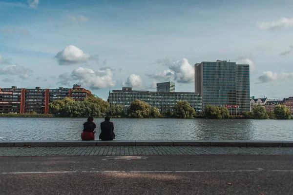 Persone sedute sul lungomare vicino al canale con edifici e cielo nuvoloso sullo sfondo, Copenaghen, Danimarca — Foto stock