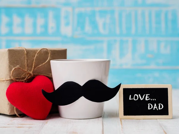 Happy father's day concept.  Gift box, a cup of coffee with mustache, red heart and wooden tag with LOVE DAD text on white and blue background.