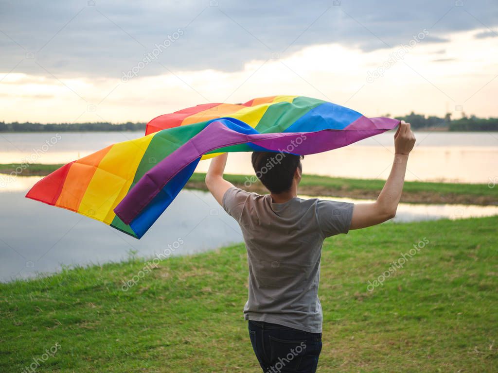 Young man holding a rainbow flag over his head beside the lake on sunset sky background. LGBT, Gay, Pride Concept.