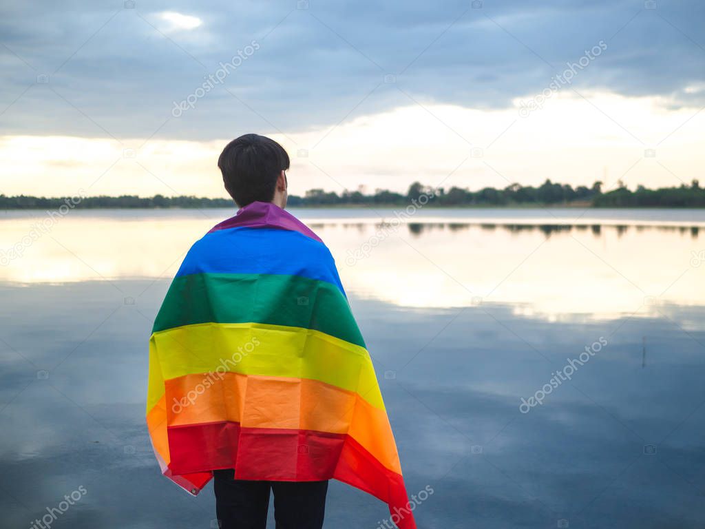Young man covered by a rainbow flag  beside the lake on sunset sky background. LGBT, Gay, Pride Concept.