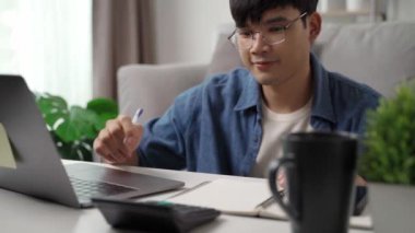 Günlük kıyafetler giyen, dizüstü bilgisayarla çalışan, akıllı telefon, hesap makinesi olan, evdeki oturma odasındaki kanepede oturan, ev konseptiyle çalışan bir adam..
