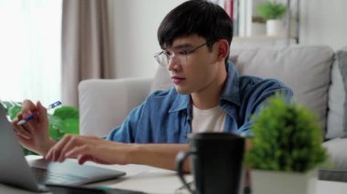 Günlük kıyafetler giyen, dizüstü bilgisayarla çalışan, akıllı telefon, hesap makinesi olan, evdeki oturma odasındaki kanepede oturan, ev konseptiyle çalışan bir adam..