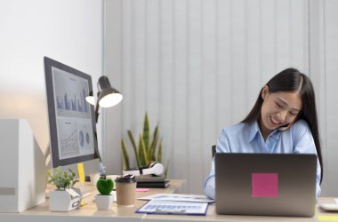 Asyalı kadın çalışan telefonda konuşuyor ve gülümseyen bir yüzle dizüstü bilgisayarda çalışıyor, ofis personeli çalışıyor ve telefon konuşmaları yapıyor..