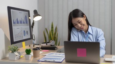 Asyalı kadın çalışan telefonda konuşuyor ve gülümseyen bir yüzle dizüstü bilgisayarda çalışıyor, ofis personeli çalışıyor ve telefon konuşmaları yapıyor..