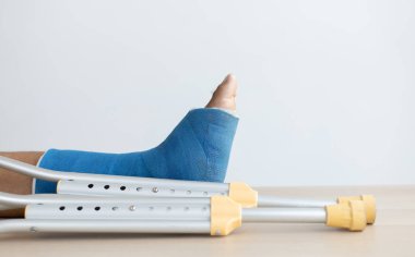 Genç bir adamın düştüğü kazadan bacağında mavi bilek ve ayak bantları, ayak bileğinde mavi alçı, koltuk değnekleri hastanın yürümesine yardımcı oluyor..