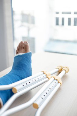 Genç bir adamın düştüğü kazadan bacağında mavi bilek ve ayak bantları, ayak bileğinde mavi alçı, koltuk değnekleri hastanın yürümesine yardımcı oluyor..