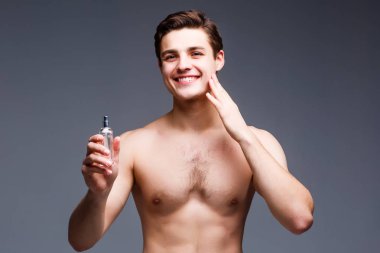 Gömleksiz genç adam gri arka plan üzerinde parfüm püskürtme