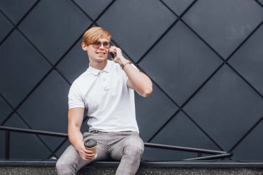 Genç sarışın adam güneş gözlüğü ve beyaz gömlek gri pantolon konuşmak smartphone ile