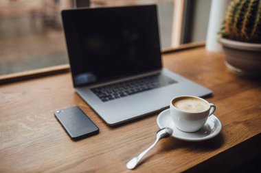 Kafede akıllı telefon ve dizüstü bilgisayar yakınında masada kaşıkla beyaz fincan kahve, seçici odak