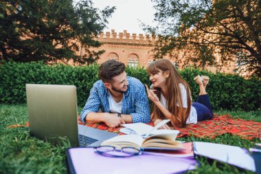 İki öğrenci dizüstü bilgisayarla çimenlerin üzerinde yatarak ödev lerini yapıyor 