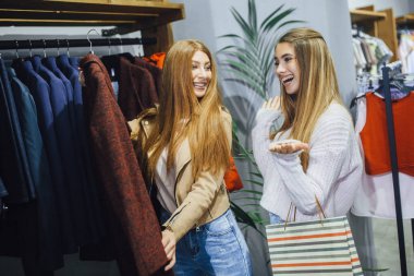 İki genç kadın ceket seçimi ve çarşıda alışveriş yaparken gülümseyen hediye çantaları ile