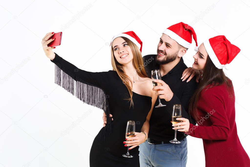 Two beautiful women and man in red christmas hats making selfie isolated on white background