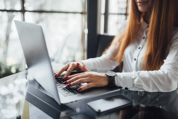 Young woman dressed in white blouse with red nails and expensive watch, typing on laptop on table