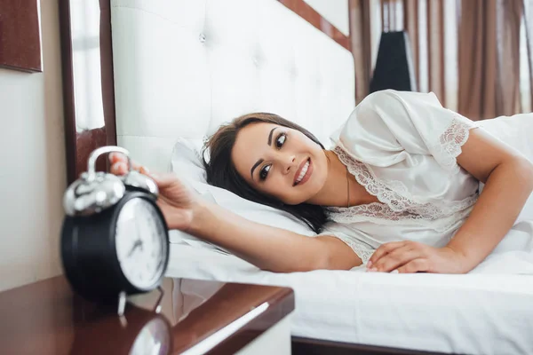 young brunette woman in morning waking up in bed with black alarm clock
