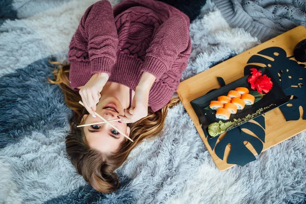 young woman lying and smiling in bed and eating sushi with chopsticks