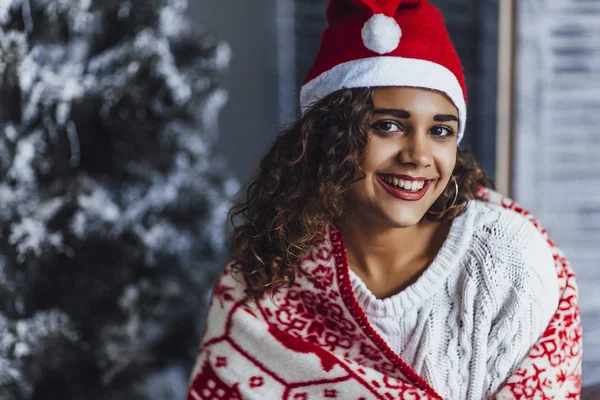 young beautiful curly woman wearing Santa Clause hat and wrapped in warm blanket, drinking hot coffee on bed in loft room