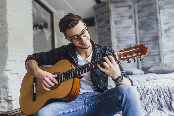 Young handsome man in glasses smiling and posing with guitar on bed 