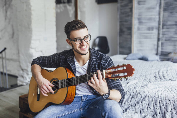 Young handsome man in glasses smiling and posing with guitar on bed 