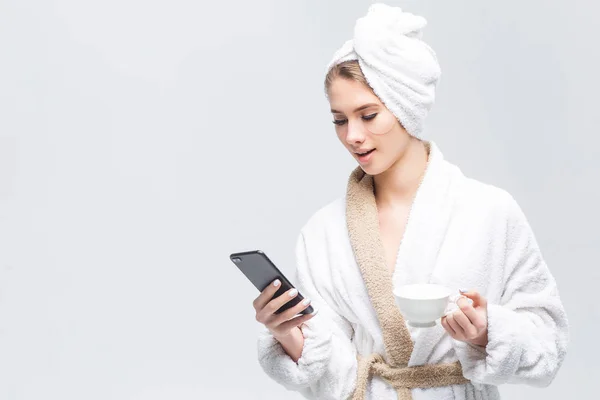 woman resting at home in robe with towel on head and using smartphone with white cup in hand isolated on white background