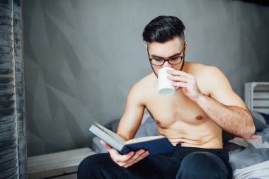 Kitap okurken yatak tutan kahve bardağı üzerinde gömleksiz döşeme yakışıklı genç adam