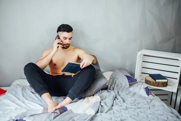 Junger Mann Liegt Mit Buch Bett Und Spricht Mit Smartphone — Stockfoto