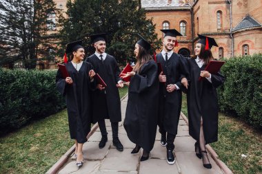 Diploma tutan akademik elbiselerde başarılı mezunlar, kameraya bakıyor 