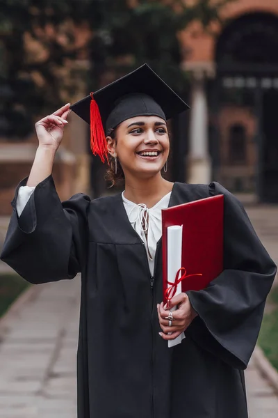 Mezuniyet önlüklerinde düşünceli genç kadın diploma tutan ve uzağa bakan