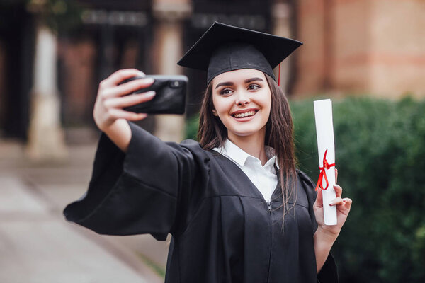 Graduated woman in graduation hat and gown making selfie