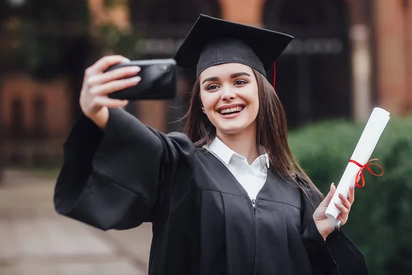 Graduated woman in graduation hat and gown outdoors making selfie