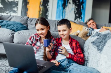 mutlu çocuklar dizüstü bilgisayarda oynarken ve çay içme, arka planda uyuyan dedesi