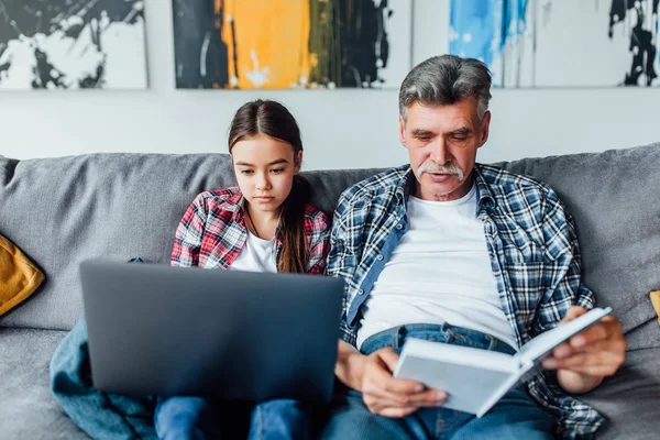 Abuelo Leyendo Libro Mientras Que Nieta Usando Ordenador Portátil —  Fotos de Stock