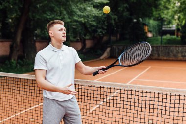 Tenis raketi taşıyan ve tenis kortunda gülümseyen polo tişörtlü genç adam.