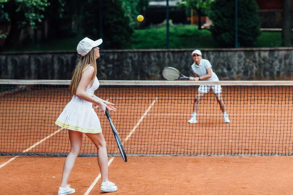 Açık tenis kortu eğitimi sırasında tenis topu servisi yapan şapkalı iki kadın ve tenis üniforması