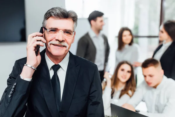 Handsome older boss talking on smartphone, colleagues in background