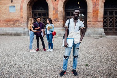 Genç, oldukça neşeli başarılı afrikalı nerdy öğrenci i, kitaplar ile ayakta, arka planda sınıf arkadaşları, kampüs yakınındaki park, güneşli bir gün .