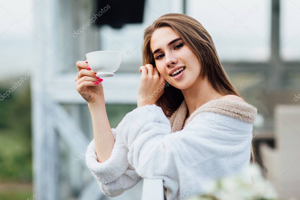 Smiling young female posing with cup of some drink, wearing at white robe.