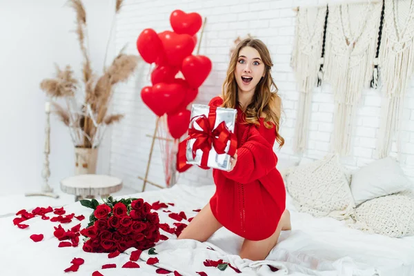 Duygusal kız hediye kutusunu gösteriyor ve yatakta sevgililer günü hediyeleriyle otururken kameraya bakıyor.