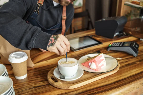Kahveci adam kahvecide latte sanatı yapıyor, masada pasta, kahveye çiçek çiziyor, yakın plan..