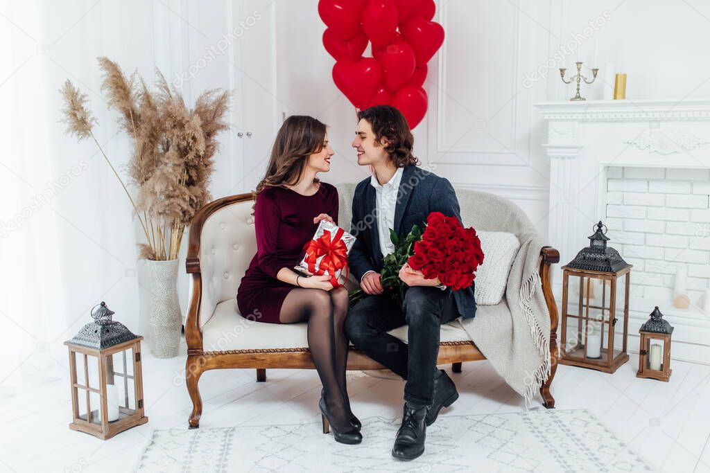 happy couple sitting on the sofa and looking at each other with love, holding flowers and present in the hands with heart shaped balloons on the background