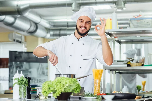 Portrait of handsome bearded man chief cook holding a saucepan and make okay sign. Profession and people concept.