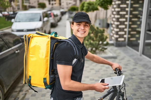 Portrait of happy delivery man in uniform on a bicycle near a modern house, with a yellow backpack.